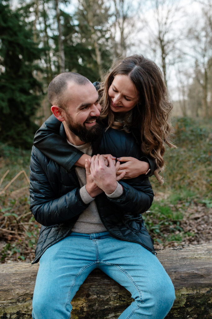 Chelsey wraps her arms around Toms chest from behind whilst he sits down on a log on the shoot in oakley wood