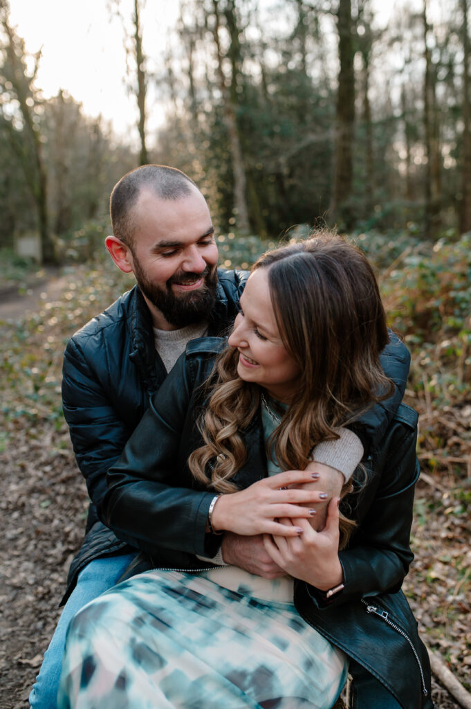 Tom wraps his arms around Chelsey whilst they both sit on the edge of a log and laugh in this shoot in oakley woods