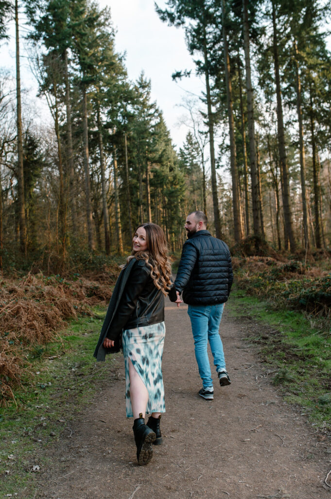 Tom and Chelsey hold hands and walk away as chelsey peers back over her left shoulder in oakley woods