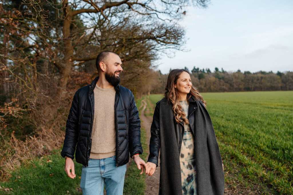 Chelsey and Tom walk hand in hand across the fields by oakley wood looking into the distance smiling