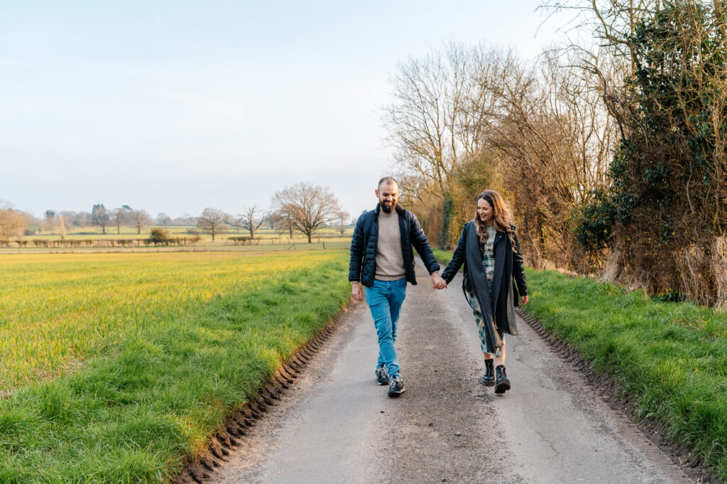 Chelsey and Tom walk down the lane holding hands by the fields next to oakley wood