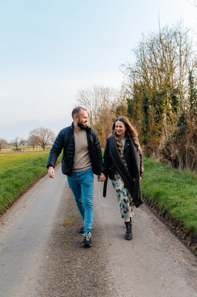 Chelsey and Tom walk down the lane holding hands looking at each other by the fields next to oakley wood
