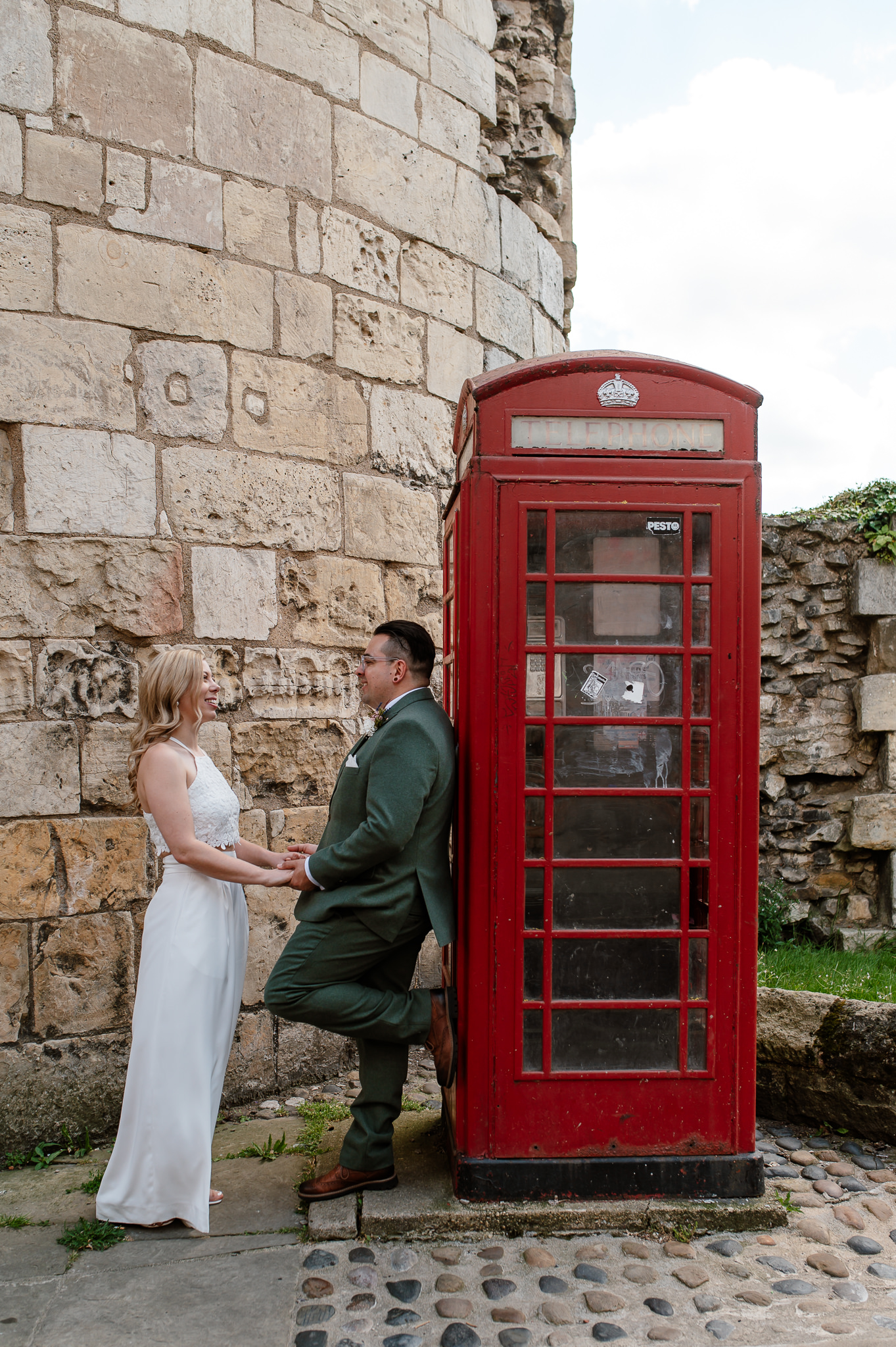 groom leaning against a telephone box whilst holding hands with bride